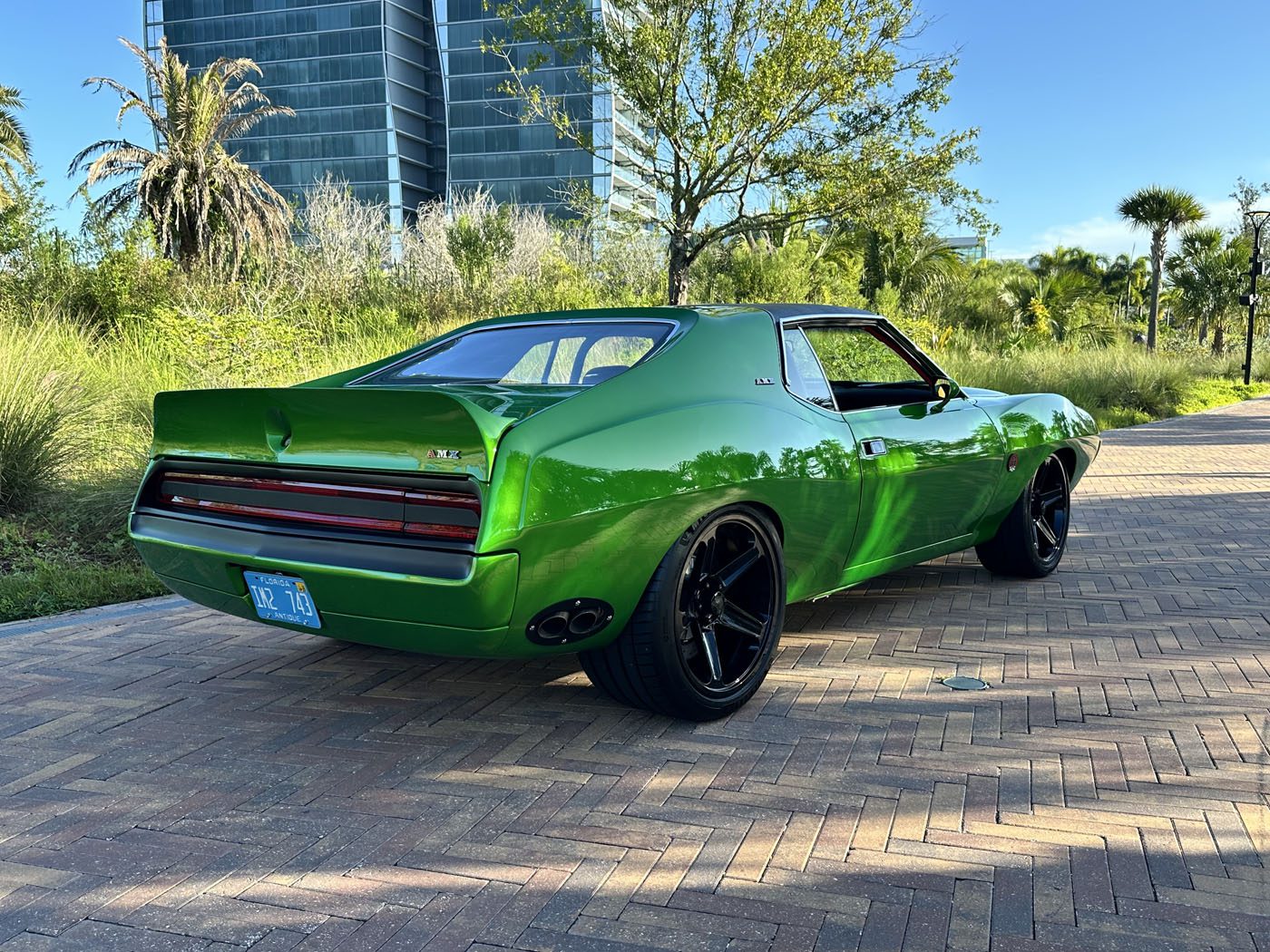 A bright green 1972 AMC Javelin muscle car with black wheels is parked on a brick road near modern buildings and lush greenery, showcasing true Pro Touring style.