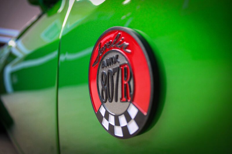 Close-up of a red and black circular "Javelin AMX 401 R" badge with a checkered pattern on the side of a green 1972 AMC Javelin, highlighting its Pro Touring style.
