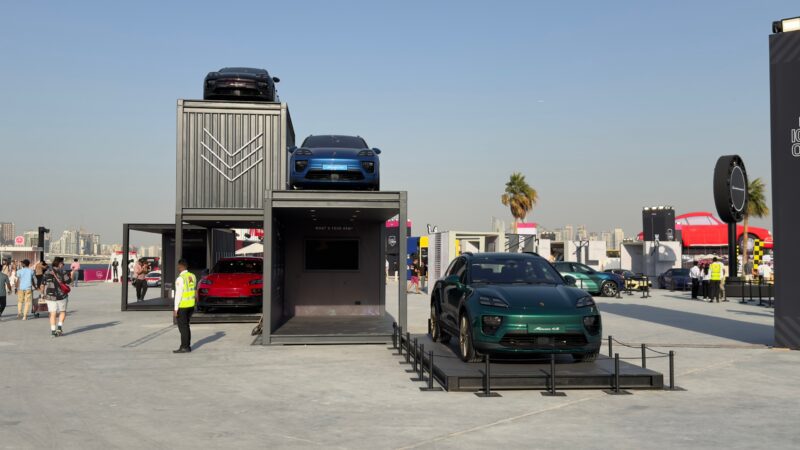 Feature Exclusive: 2025 Icons of Porsche Dubai showcases several sports cars displayed on and inside stacked shipping containers at an outdoor auto show, with people walking nearby under a clear sky.