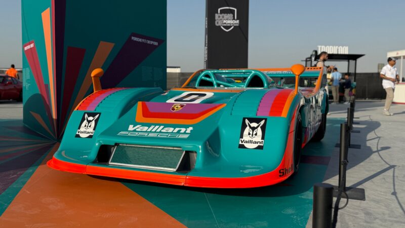 Feature Exclusive: A teal and orange Vaillant Porsche race car on display outdoors, surrounded by barriers and informational signage at the 2025 Icons of Porsche Dubai.
