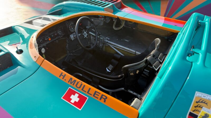 Cockpit of a teal and orange race car with the name "H. Müller" and a Swiss flag decal on the side. Racing seat, steering wheel, and dashboard are visible. Feature Exclusive: 2025 Icons of Porsche Dubai.