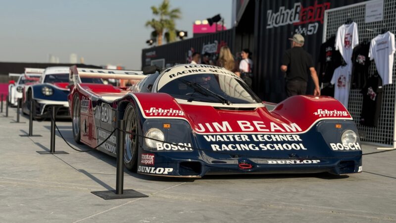 Feature Exclusive: 2025 Icons of Porsche Dubai—a vintage Jim Beam Walter Lechner Racing School race car is displayed outdoors, with merchandise and people in the background.