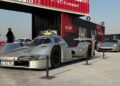 Feature Exclusive: 2025 Icons of Porsche Dubai—a silver Porsche race car and a silver sports car are displayed outdoors on a concrete surface, with people and shipping container structures in the background.
