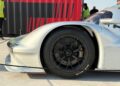 Feature Exclusive: Close-up of the front left side of a silver race car with a black Michelin tire, parked near a red and black striped background at the 2025 Icons of Porsche Dubai, with people in safety vests standing nearby.