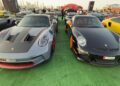 Feature Exclusive: 2025 Icons of Porsche Dubai—two stunning Porsche sports cars, one grey with red accents and one black with orange accents, parked side by side at an outdoor car event amid an array of impressive vehicles.