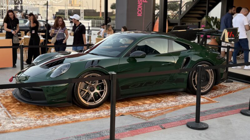 Feature Exclusive: A dark green Porsche 911 GT3 RS is displayed on a rug behind barriers at the 2025 Icons of Porsche Dubai outdoor event, with people standing and walking nearby.