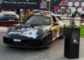 A custom sports car with colorful graffiti art is displayed outdoors at the Feature Exclusive: 2025 Icons of Porsche Dubai, featuring cartoon designs and an open top, with a sign and crowd in the background.
