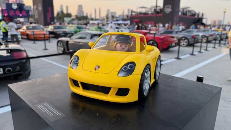 Feature Exclusive: 2025 Icons of Porsche Dubai—a yellow toy Porsche with a Playmobil driver sits on a black platform at an outdoor car show, set against luxury cars and the dazzling city skyline.
