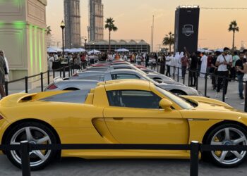 Feature Exclusive: 2025 Icons of Porsche Dubai—a row of sports cars, led by a yellow Porsche Carrera GT, is displayed at an outdoor event attended by a crowd of people.
