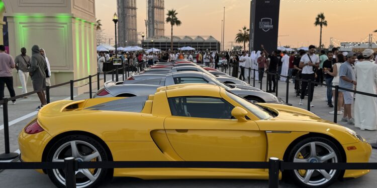 Feature Exclusive: 2025 Icons of Porsche Dubai—a row of sports cars, led by a yellow Porsche Carrera GT, is displayed at an outdoor event attended by a crowd of people.