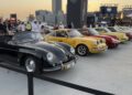 A row of classic Porsche cars on display at an outdoor event, with people walking around and a city skyline in the background. Feature Exclusive: 2025 Icons of Porsche Dubai.