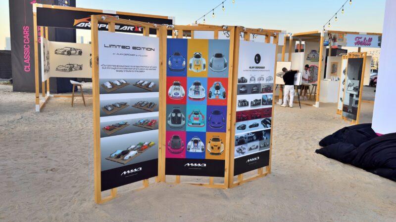 Three display panels at the sandy outdoor exhibition feature exclusive: 2025 Icons of Porsche Dubai model cars, with various open wooden booths and people visible in the background.