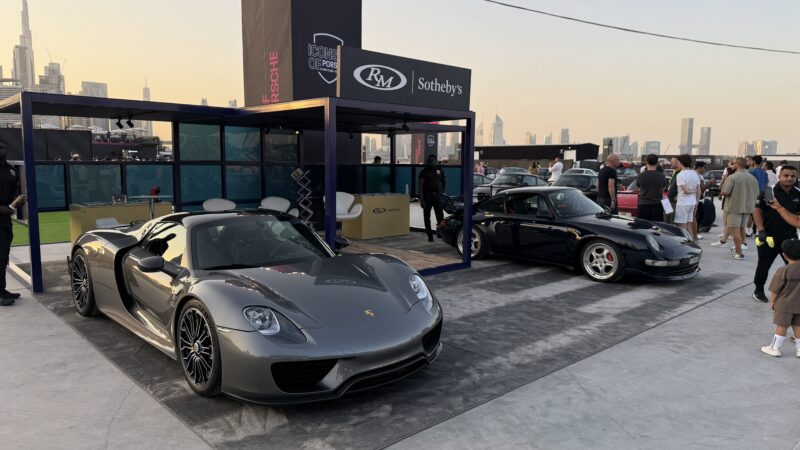 Feature Exclusive: Two Porsche sports cars are displayed outdoors at an RM Sotheby’s event, part of the 2025 Icons of Porsche Dubai, with people and city skyscrapers visible in the background.