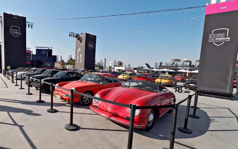 Feature Exclusive: 2025 Icons of Porsche Dubai showcases a stunning display of classic and modern Porsche cars at an outdoor event, with barriers around the vehicles and the city skyline visible in the background.
