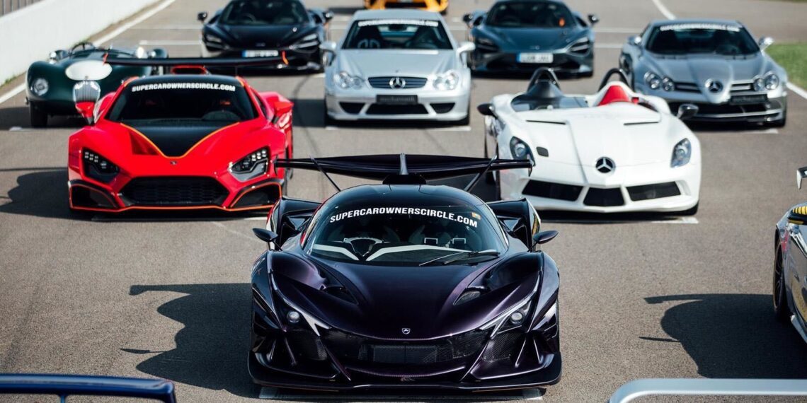 A lineup of high-performance supercars, including a black hypercar in front, parked on a racetrack under clear daylight.