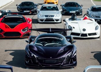 A lineup of high-performance supercars, including a black hypercar in front, parked on a racetrack under clear daylight.