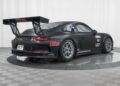 A matte black Porsche 911 GT3 race-ready car with a large rear wing, racing decals, and number 18, displayed in a studio with a checkered floor and white background. Offered by Isringhausen Imports for true Porsche track car enthusiasts.