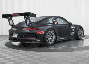 A matte black Porsche 911 GT3 race-ready car with a large rear wing, racing decals, and number 18, displayed in a studio with a checkered floor and white background. Offered by Isringhausen Imports for true Porsche track car enthusiasts.