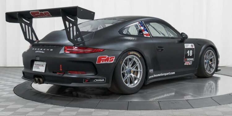 A matte black Porsche 911 GT3 race-ready car with a large rear wing, racing decals, and number 18, displayed in a studio with a checkered floor and white background. Offered by Isringhausen Imports for true Porsche track car enthusiasts.