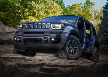 A trail-ready 2026 Jeep Recon, all-electric and dark gray, with no doors or roof panels, is parked on rocky terrain surrounded by trees and greenery under a clear sky.