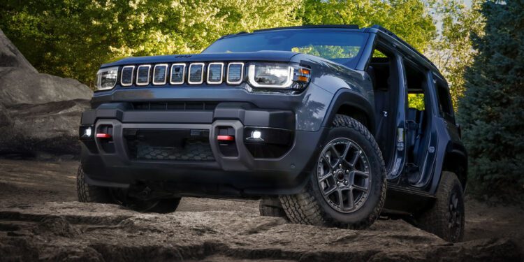 A trail-ready 2026 Jeep Recon, all-electric and dark gray, with no doors or roof panels, is parked on rocky terrain surrounded by trees and greenery under a clear sky.
