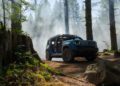 A blue, trail-ready Jeep SUV with open doors is parked on a dirt clearing in a forest, surrounded by trees and mist.