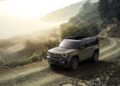 A Trail-Ready, all-electric 2026 Jeep Recon navigates a winding dirt road through a mountainous, misty landscape with trees and rugged terrain.