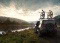 A trail-ready 2026 Jeep Recon, all-electric and adventure-ready, is parked by a riverbank with two bicycles mounted on the roof, surrounded by trees and hills under a cloudy sky.