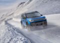 A trail-ready, all-electric 2026 Jeep Recon drives on a snow-covered mountain road, surrounded by snowbanks and snowy hills under a cloudy sky.