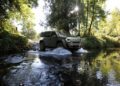 A trail-ready 2026 Jeep Recon, an all-electric SUV, drives through a shallow creek surrounded by trees and sunlight filtering through the foliage.
