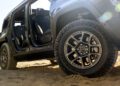 Close-up of the trail-ready 2026 Jeep Recon, an all-electric SUV parked on sand with its doors removed, highlighting rugged tires and interior seating in daylight.