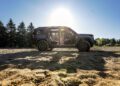 A trail-ready, all-electric 2026 Jeep Recon with open doors is parked on sandy ground in front of trees, illuminated by bright sunlight streaming from behind.