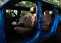 A blue, all-electric 2026 Jeep Recon with its doors removed reveals brown leather front seats and a sunlit interior, trail-ready and parked in a wooded area.