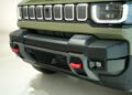 Close-up of the front bumper and grille of the 2026 Jeep Recon, an all-electric Jeep, in olive green with red tow hooks and LED headlights—highlighting its bold, trail-ready design.