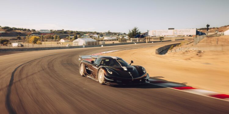 A black Koenigsegg Sadair’s Spear speeds around a curve on a sunny racetrack, shattering the Laguna Seca production car record for 2025, with barriers, signage, and hills visible in the background.