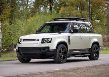 A white Defender Pickup with black wheels and a roof rack from Heritage Customs is parked on a paved road, surrounded by greenery and trees.