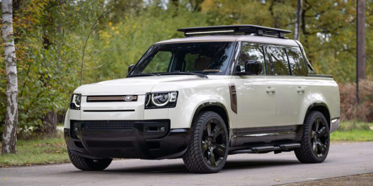 A white Defender Pickup with black wheels and a roof rack from Heritage Customs is parked on a paved road, surrounded by greenery and trees.