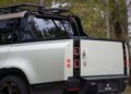 A close-up of the rear and side of a light-colored Defender Pickup with a black roof and trim, parked outdoors in a wooded area, showcases distinctive Urban Automotive styling.