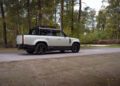 A white Defender Pickup with black wheels and roof rack is parked on a paved road, surrounded by trees and greenery—a true Urban Automotive statement blending rugged style with Heritage Customs design.