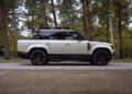 A white and black Defender Pickup, customized by Heritage Customs, is parked on a forest road, surrounded by tall trees and greenery.