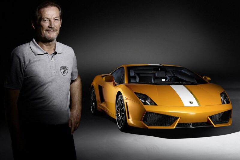 Vyras pilkais Lamborghini polo marškinėliais stovi prie oranžinės spalvos sportinio automobilio su balta lenktynine juostele – paskutinio Lamborghini su galinių ratų pavara ir mechanine pavarų dėže, pastatyto tamsiame fone.