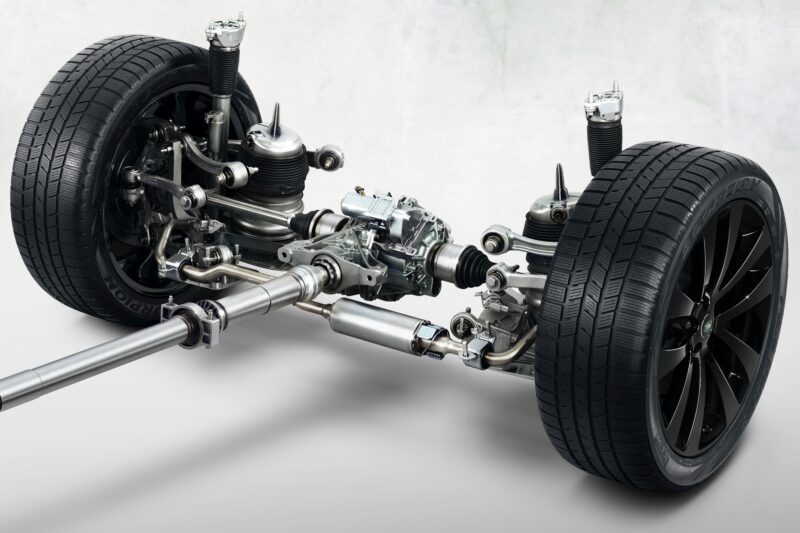 A close-up view of the 2025 Land Rover Range Rover Sport Dynamic SE PHEV's rear suspension and drivetrain components, showcasing wheels, shock absorbers, axles, and connecting rods against a plain background.