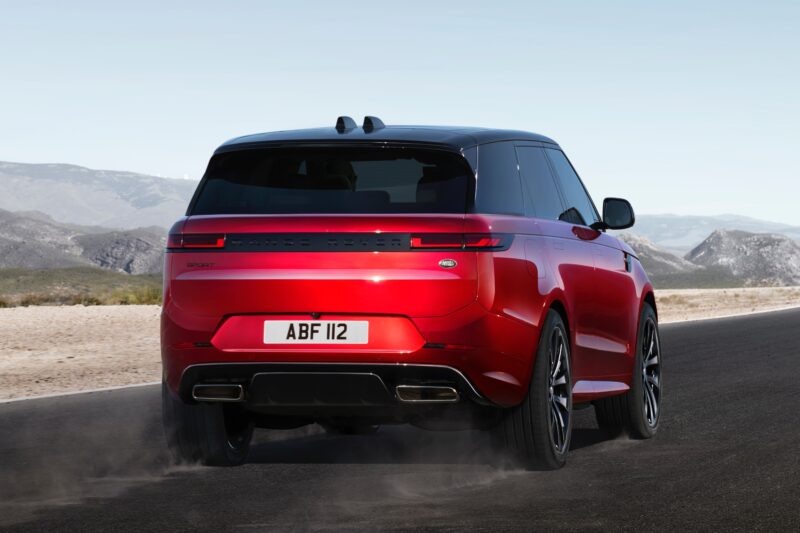 A red Range Rover SUV, featured in the Review: 2025 Land Rover Range Rover Sport Dynamic SE PHEV, drives on a paved road in a desert landscape, viewed from behind with dust trailing from the tires.