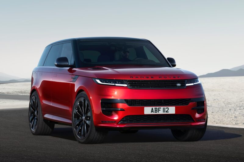 A red 2025 Land Rover Range Rover Sport Dynamic SE PHEV drives on a paved road through a desert landscape under a clear sky.
