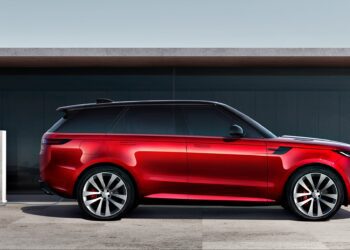 A red SUV, the 2025 Land Rover Range Rover Sport Dynamic SE PHEV, is parked next to an electric vehicle charging station on a concrete surface, with a modern building in the background.