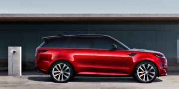 A red SUV, the 2025 Land Rover Range Rover Sport Dynamic SE PHEV, is parked next to an electric vehicle charging station on a concrete surface, with a modern building in the background.
