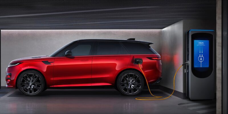 A red SUV, the 2025 Land Rover Range Rover Sport Dynamic SE PHEV, is parked in a garage and connected to an electric vehicle charging station via a yellow cable.