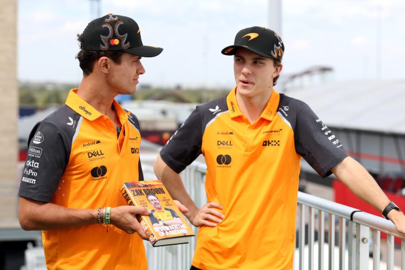 Two people in orange and black team shirts and caps stand on a balcony, one holding "Zak Brown: Surviving to Thriving," as they discuss how McLaren Racing CEO Zak Brown reveals secrets to success in his new autobiography.