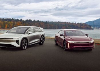 Two modern electric cars, one silver and one maroon, parked side by side on a paved area with a scenic lake, bridge, and autumn trees—reminiscent of Lucid's record Q3 deliveries amid the EV market slowdown.