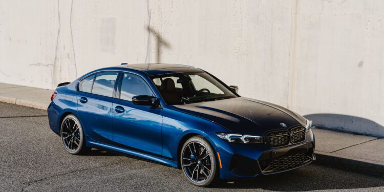 A blue BMW M340i xDrive sedan, 50 Jahre Edition, is parked on a sunlit street next to a light-colored concrete wall.